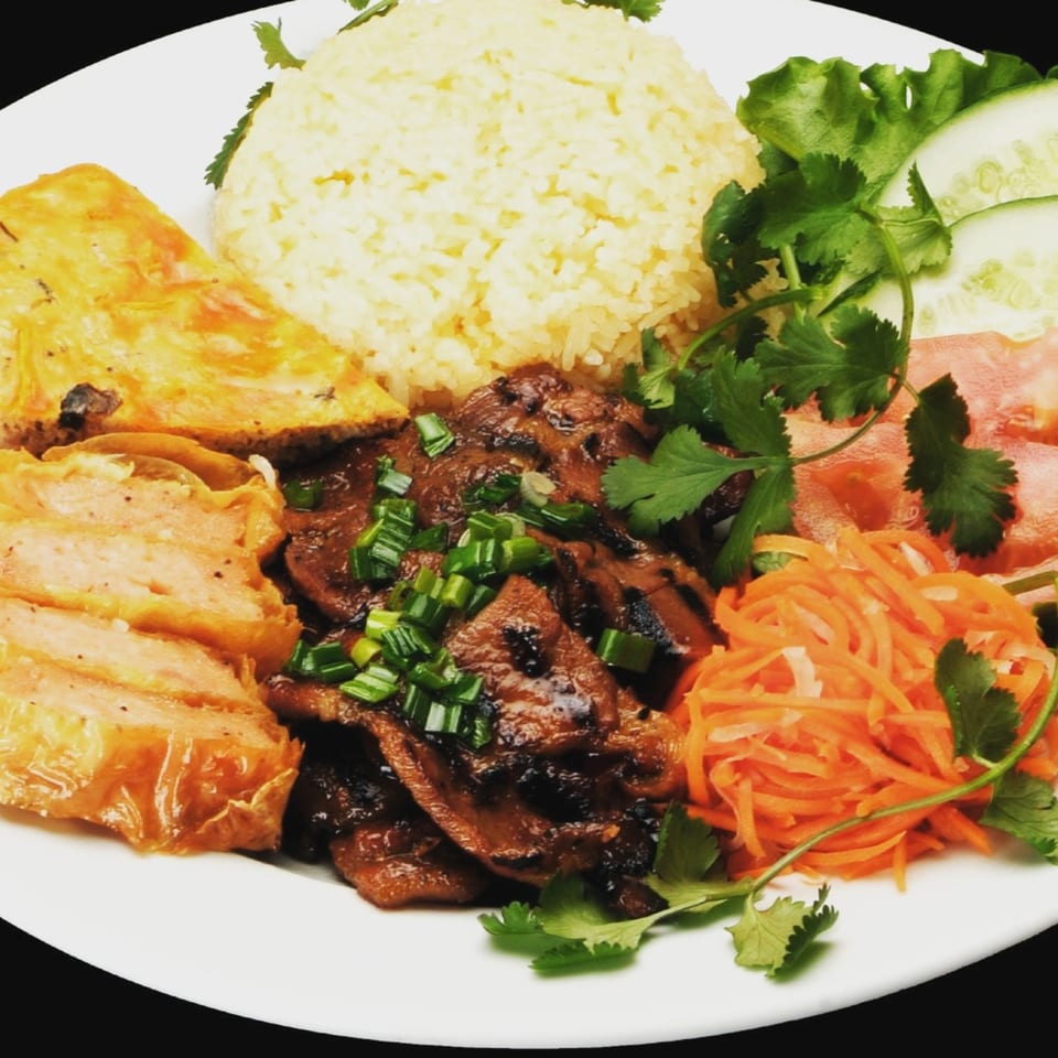 Vietnamese grilled pork, fried egg cake, rice, pickled carrots, and fresh vegetables on a white plate.