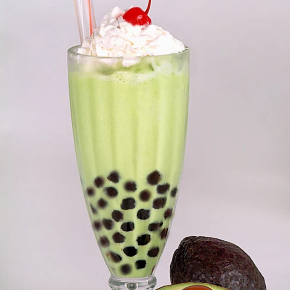 Avocado boba smoothie with whipped cream and a cherry, next to whole and halved avocados.