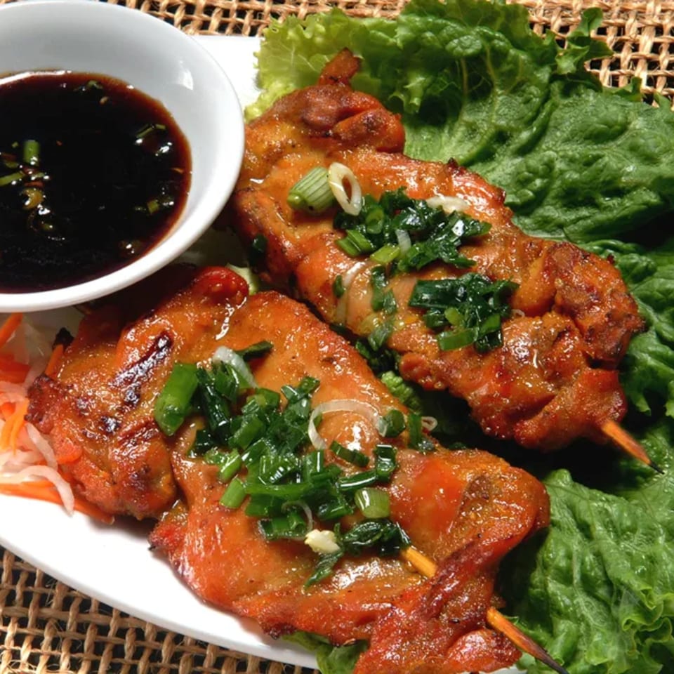 Two Vietnamese grilled chicken skewers with green onions, lettuce, and a side of dark sauce.
