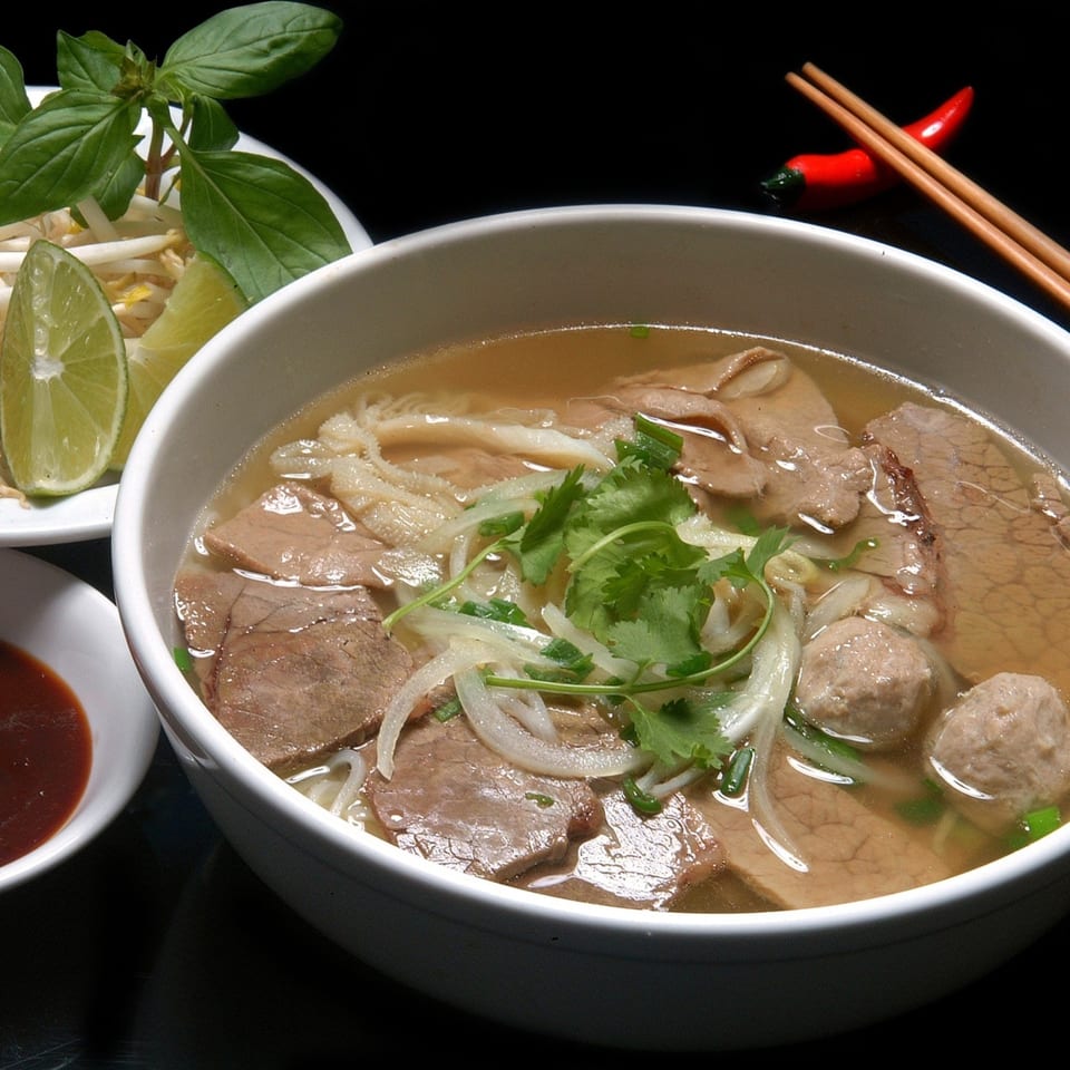 A bowl of Vietnamese pho with beef, tripe, meatballs, and fresh herbs, served with lime and bean sprouts.